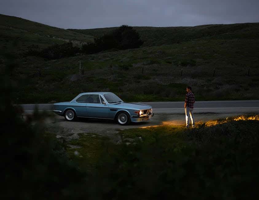 A person stands beside a vintage blue car parked on the side of a desolate road, surrounded by rolling green hills under a dim, overcast sky. The car's headlights illuminate the ground in front, adding a soft glow to the scene.