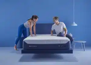 A man and woman are setting up a bed in a modern, minimalistic bedroom with blue walls and floor. The woman is on the left side of the bed, while the man is on the right side. The bed has a white mattress and a blue frame. A white hanging lamp is on the right.