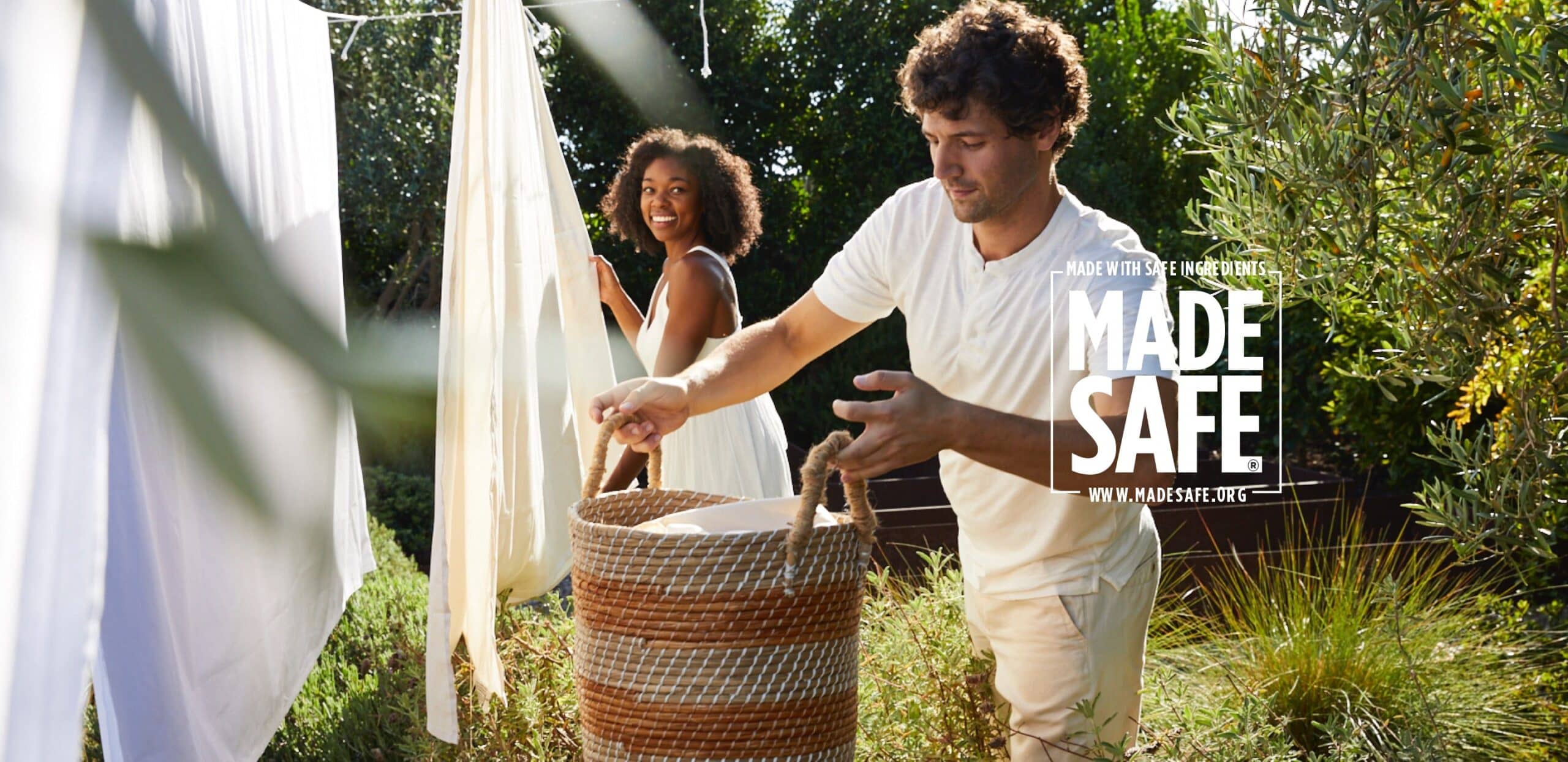A man places laundry into a large wicker basket while a smiling woman hangs white sheets on a clothesline. The background shows lush greenery. Text overlay reads: "MADE SAFE - Made with Safe Ingredients. www.madesafe.org".