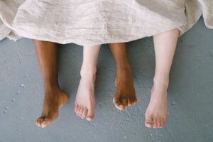 Four feet of two individuals, one with darker skin and the other with lighter skin, are peeking out from under a beige blanket. The floor beneath them is a textured gray surface. The image conveys a sense of togetherness and relaxation.