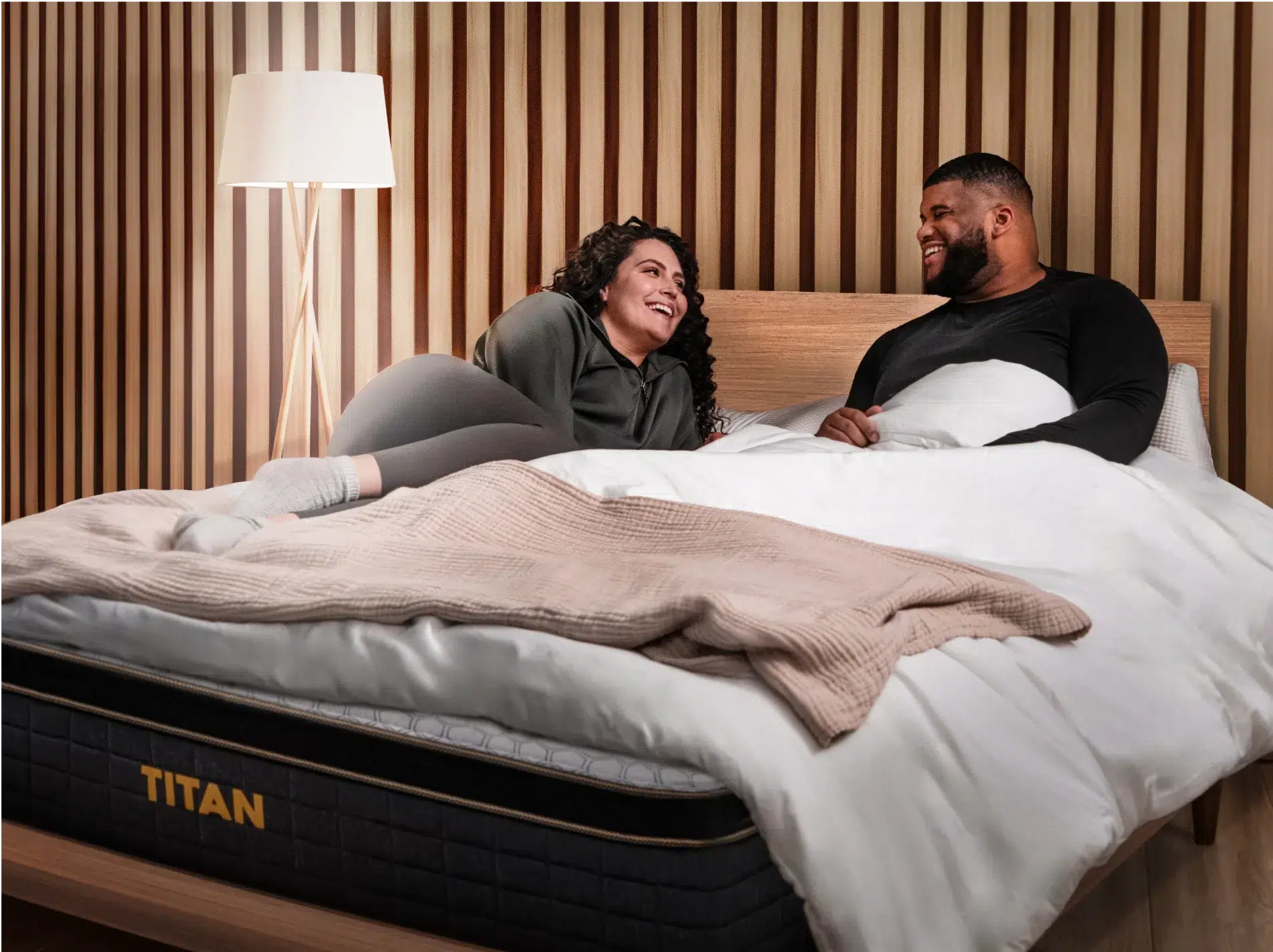 A woman and a man are lying on a bed in a cozy room with wooden paneling on the walls and a lamp in the background. They are smiling and looking at each other. The bed, labeled "Titan Plus Elite," is neatly made with white bedding, and a blanket is draped over the foot.