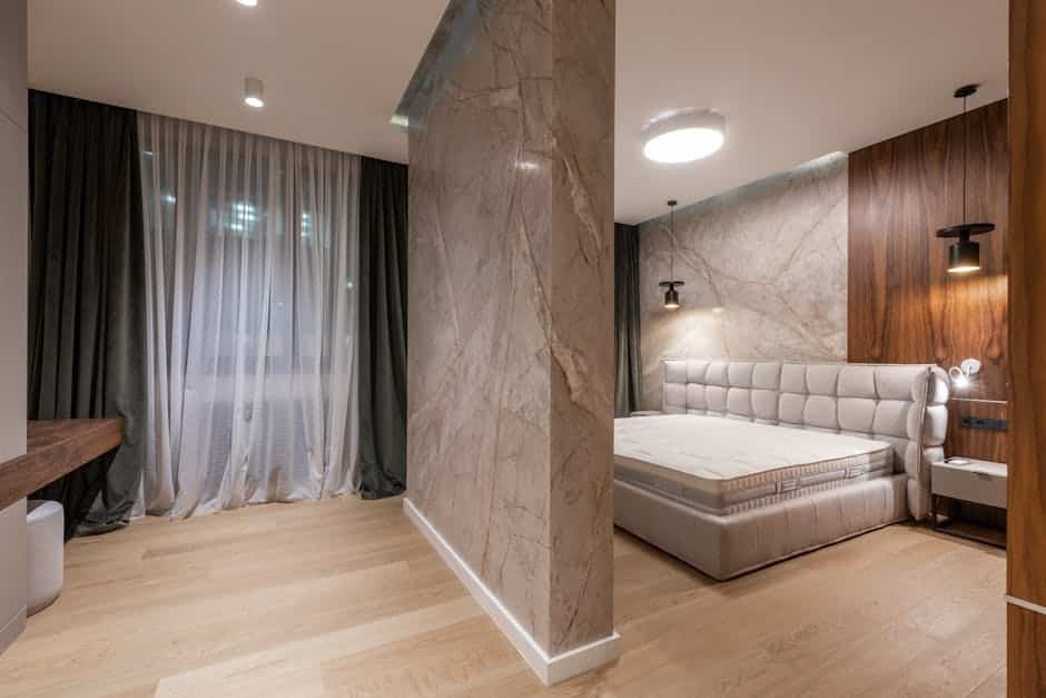 A modern bedroom with a wooden floor and a large bed with a tufted headboard. The room is divided by a marble accent wall, and features floor-to-ceiling dark curtains covering a window on the left side. Two pendant lights hang on either side of the bed.