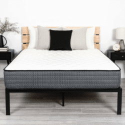 A neatly made bed showcasing the 40 Winks Studio Luxury Firm mattress and a black bed frame is flanked by two black nightstands. Each nightstand holds a lamp; the left one also has a vase with flowers, while the right one features an alarm clock. The background wall is plain white.