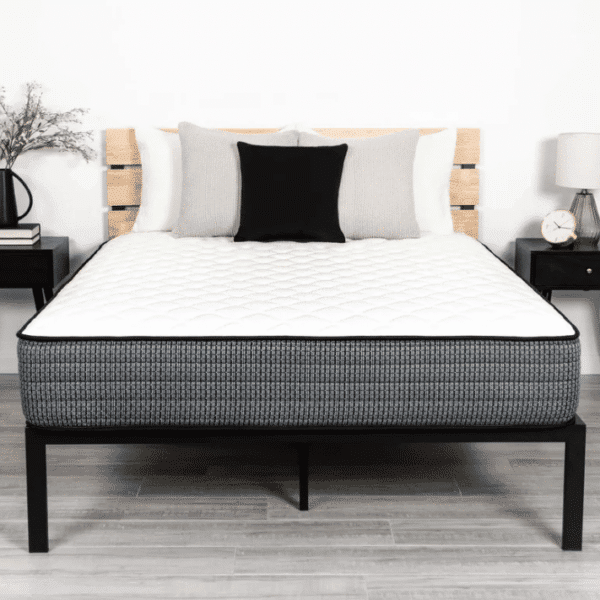 A neatly made bed showcasing the 40 Winks Studio Luxury Firm mattress and a black bed frame is flanked by two black nightstands. Each nightstand holds a lamp; the left one also has a vase with flowers, while the right one features an alarm clock. The background wall is plain white.