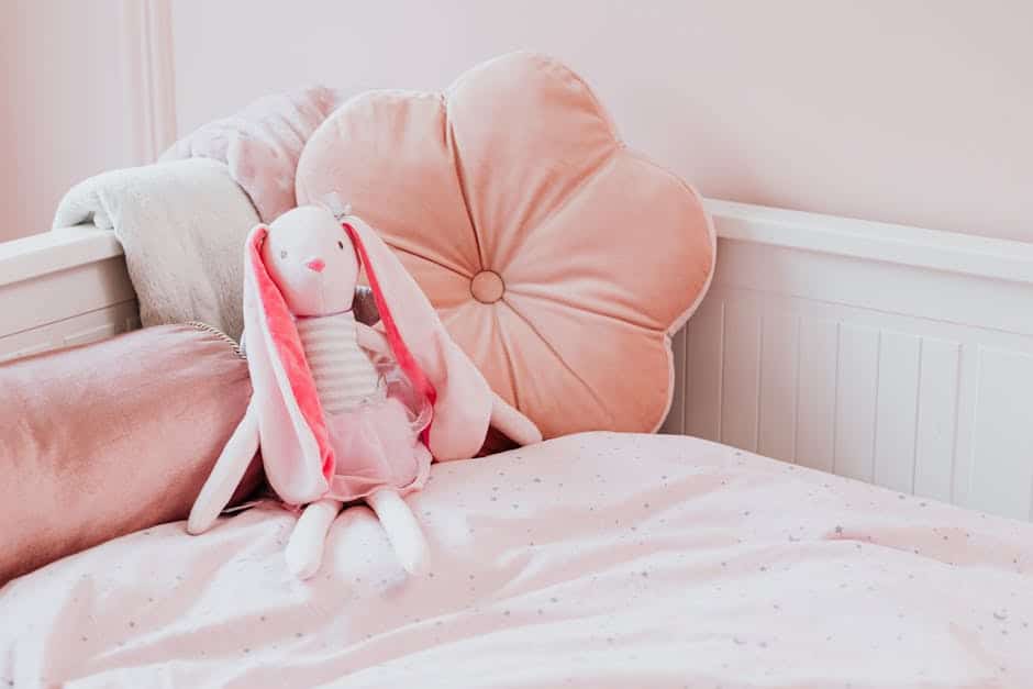 A cozy bedroom with soft pink decor featuring a plush bed with pale pink sheets, a flower-shaped cushion, and a cute stuffed bunny toy sitting upright. The stuffed bunny has long ears and is wearing a dress.