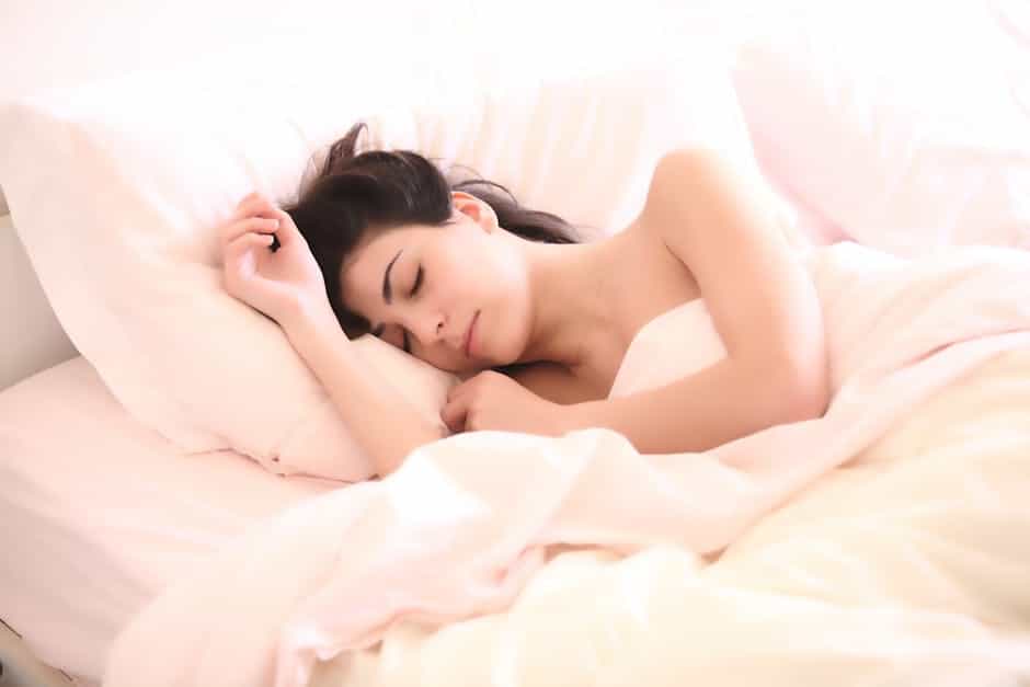 A woman with long dark hair is peacefully sleeping in a bed with white sheets and light pink blankets. One arm is resting near her head on a pillow, and she appears relaxed and comfortable in a bright, softly lit room.