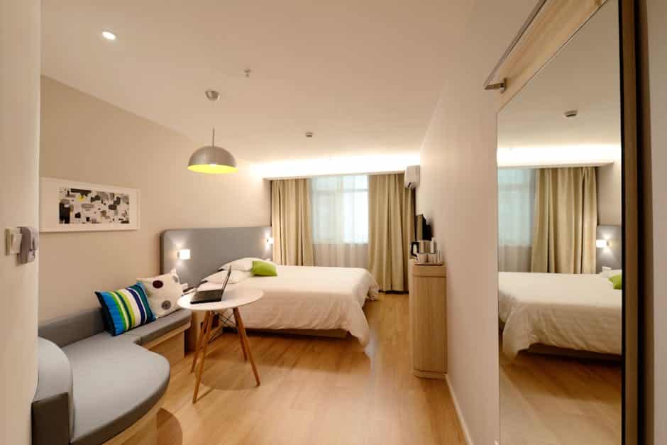 A modern hotel room with two beds, a seating area with a round table and a cushioned bench, a large mirror, a flat-screen TV, and light wood flooring. The room is well-lit with a combination of natural light from large windows and ceiling lights.