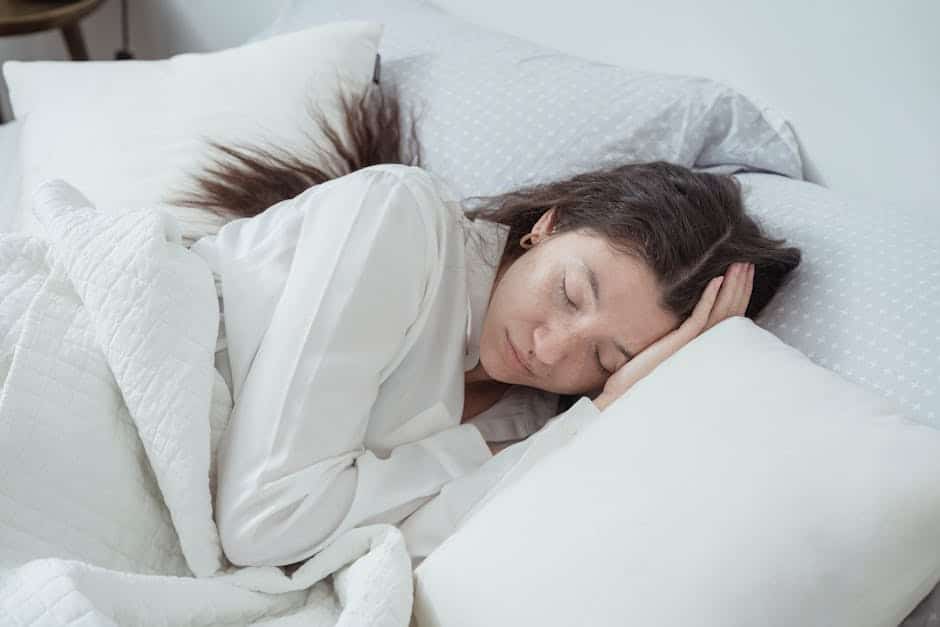 A person with long dark hair is sleeping peacefully in bed. They are lying on their side, head resting on one hand, covered by a white quilt. The bed is adorned with white pillows and light grey sheets with white polka dots.