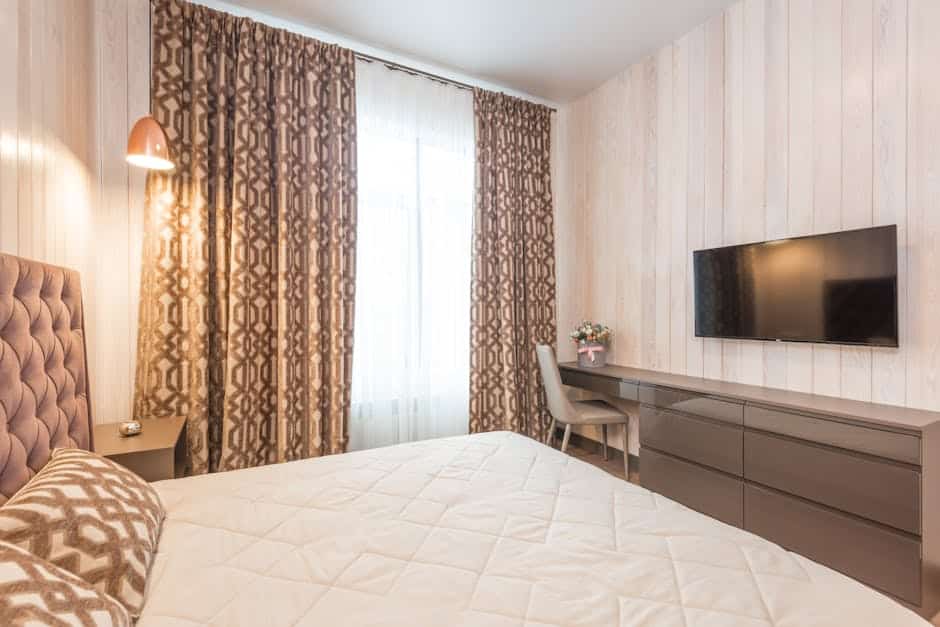 A cozy bedroom featuring a double bed with a patterned comforter, a bedside nightstand with a lamp, a large window with patterned curtains, a wall-mounted flat-screen TV, a dresser, and a chair with a small flower vase. The walls are light wood-paneled.
