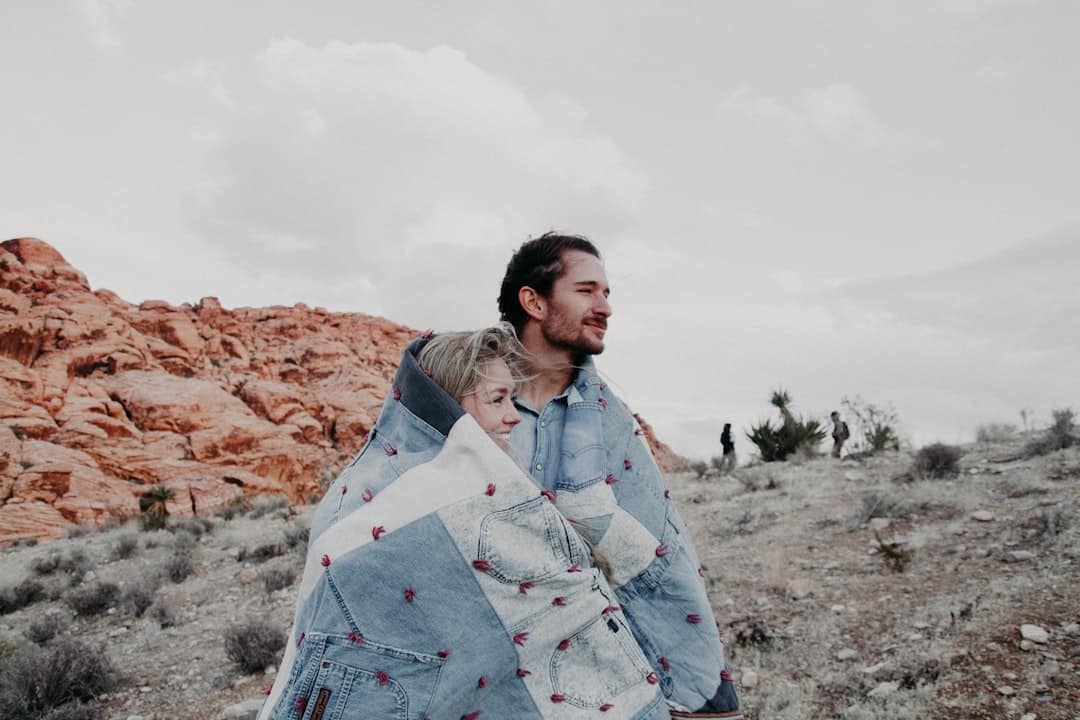 A couple stands together wrapped in a quilted blanket in a rocky desert landscape. The sky is cloudy, and they appear to be enjoying the view. Red rock formations and sparse vegetation surround them.