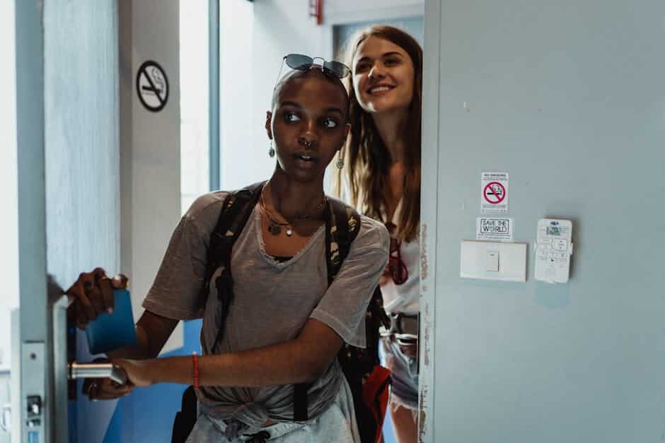 Two people entering a room. One is holding the door with a blue card in hand, wearing a gray t-shirt and backpack. The other, smiling, follows closely. There are no smoking signs on the wall beside them.