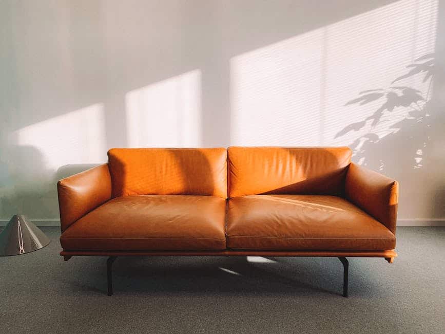 A brown leather sofa sits in a well-lit room, casting soft shadows on the wall. Sunlight streams through the window blinds, and a potted plant's silhouette is visible in the corner. A modern lamp stands to the left.