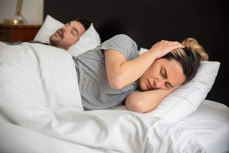 A woman in bed covers her ears with her hands, appearing frustrated, while a man sleeps next to her with his mouth open, suggesting he might be snoring. The room is softly lit, and they are under a white blanket.