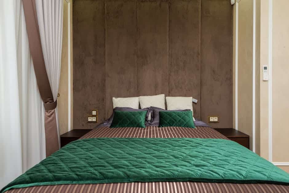 A neatly made bed with a green quilted comforter and multiple pillows is positioned against a textured brown headboard. The room features light-colored walls and a bedside table on each side of the bed. A curtain partially covers one side of the headboard.