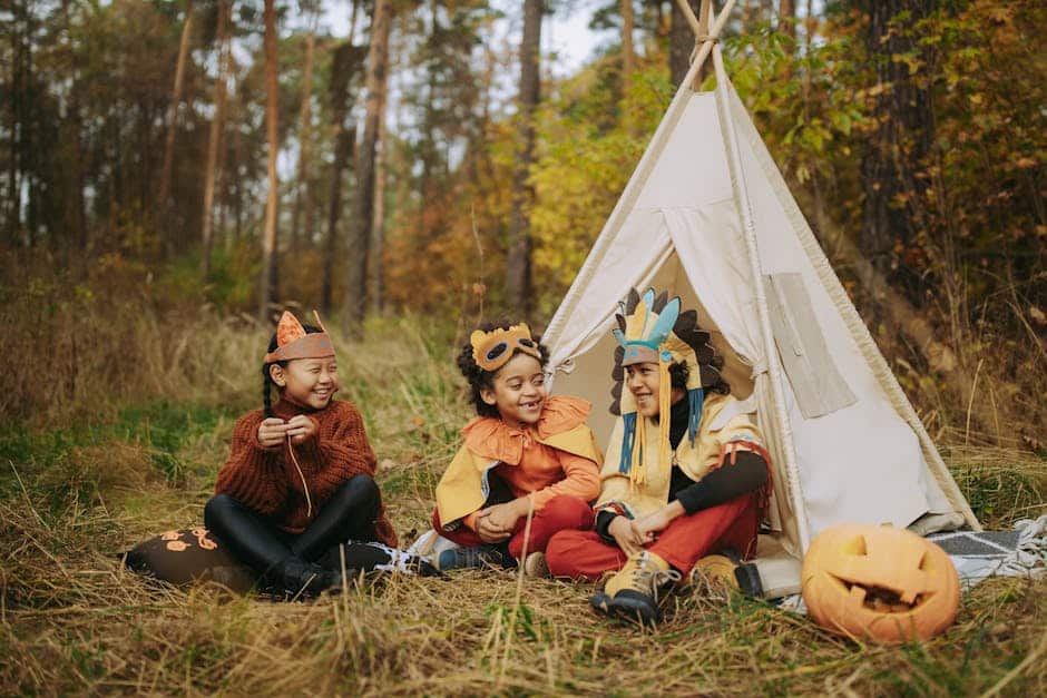Three children in animal and festive costumes sit on grass next to a small white teepee tent in a forest. They are smiling and holding props, with an illuminated carved pumpkin nearby. Tall trees and autumn leaves surround them.