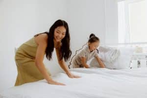 A woman and a child are making a bed together in a bright room. They are smoothing out a white bedspread. The room has white walls and a window that lets in natural light. Pillows are stacked at the head of the bed.