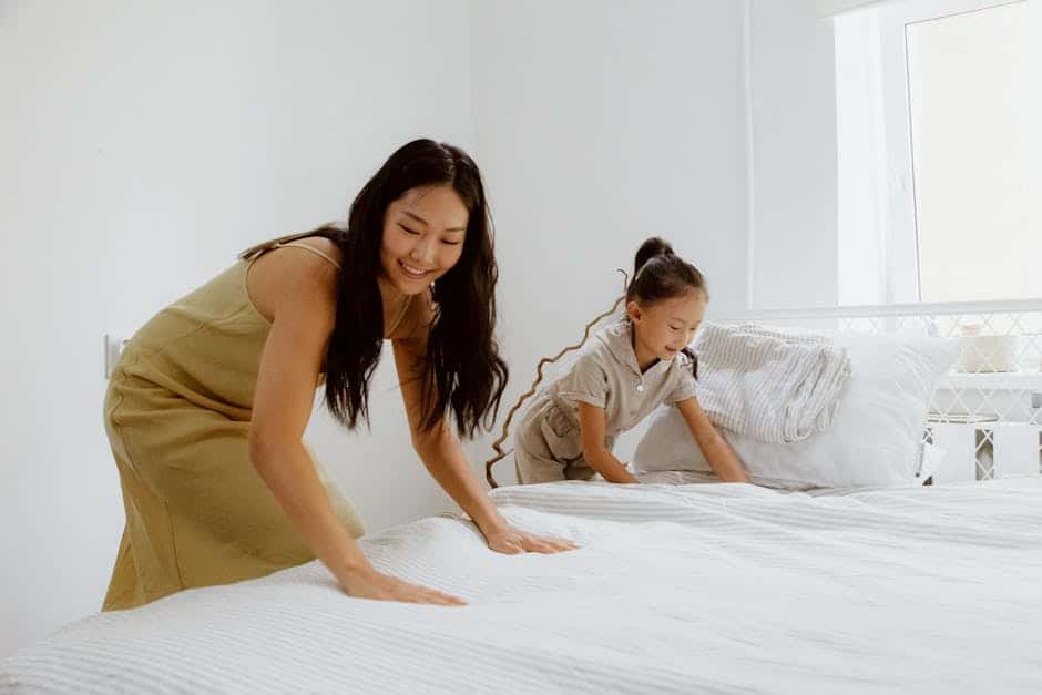 A woman and a child are making a bed together in a bright room. They are smoothing out a white bedspread. The room has white walls and a window that lets in natural light. Pillows are stacked at the head of the bed.