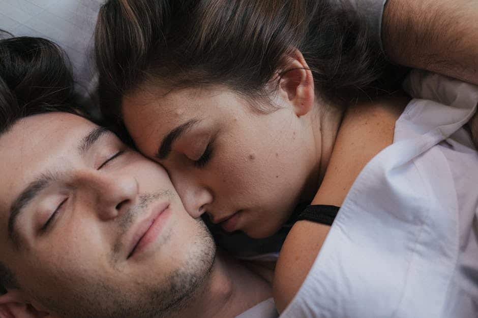 A couple peacefully sleeping, cuddled close under a white blanket. The person on the left has short dark hair and closed eyes, while the person on the right has long brown hair resting against the other's shoulder. The mood is calm and intimate.