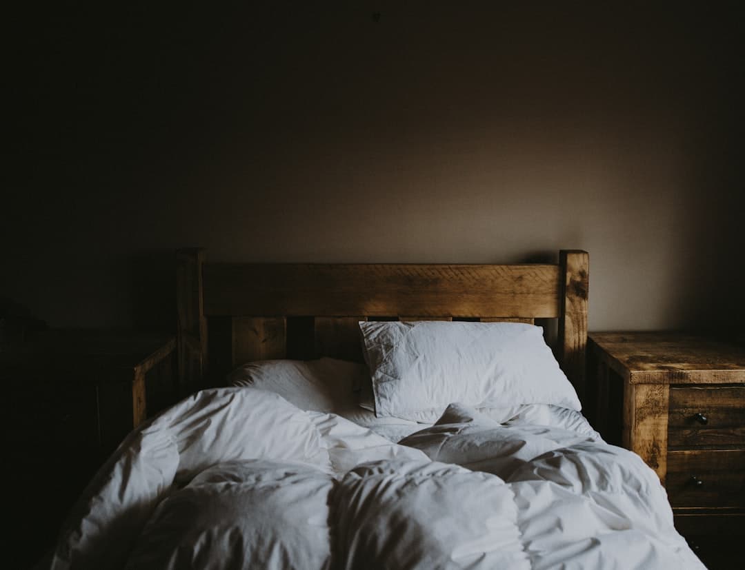 A rustic bedroom scene with a wooden bed frame. The bed is unmade, featuring white pillows and crumpled sheets. On either side of the bed are wooden nightstands, creating a warm and cozy atmosphere. The room appears dimly lit.